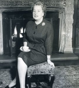 A black and white photo of Eleanor Ford sitting on an ottoman in front of a fireplace, holding a tea cup.