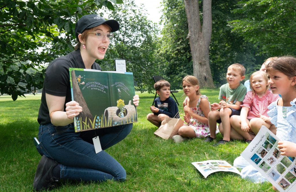 A storyteller reads a book to a group of children outside.