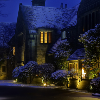 A historic stone mansion at night, softly illuminated with warm golden lights against the deep blue evening sky. Snow lightly covers the roof, trees, and bushes, creating a serene winter atmosphere. Light shines through the leaded glass windows, and a lantern above the arched entrance casts a welcoming glow.