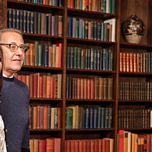 Two guests view the historic library room inside the Main Residence at Ford House, featuring rich wood paneling, shelves filled with vintage books, and a display table with a framed family photo, decorative chest, and ornate lamp.