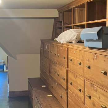 A well-organized storage room with rows of labeled wooden drawers on the right and metal shelving units holding archival boxes and rolled textiles on the left. Light from a window at the end of the room illuminates the polished floor.