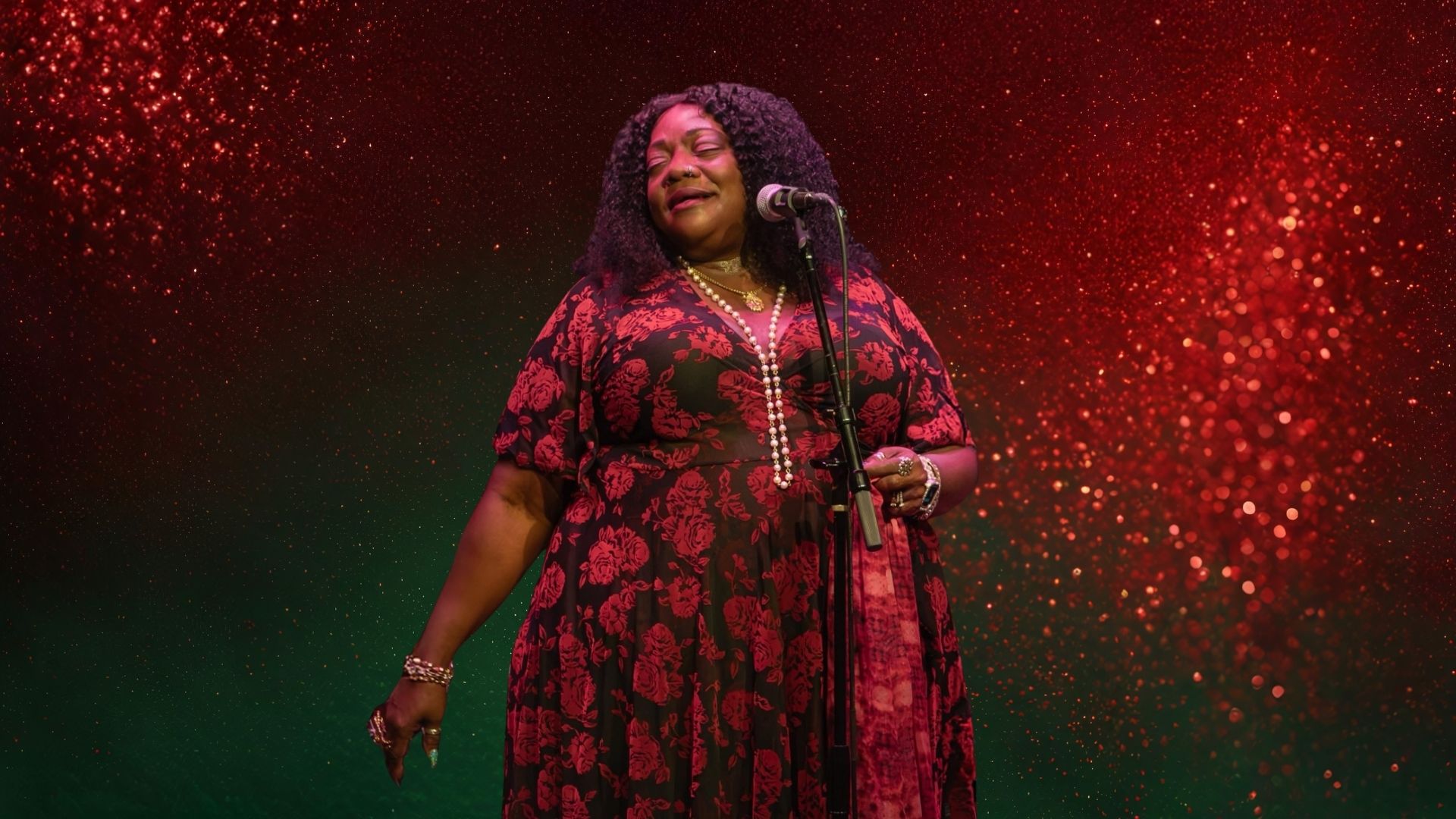 Thornetta Davis singing with eyes closed at a microphone, wearing a red floral dress and pearl necklaces, against a red and green glitter backdrop.