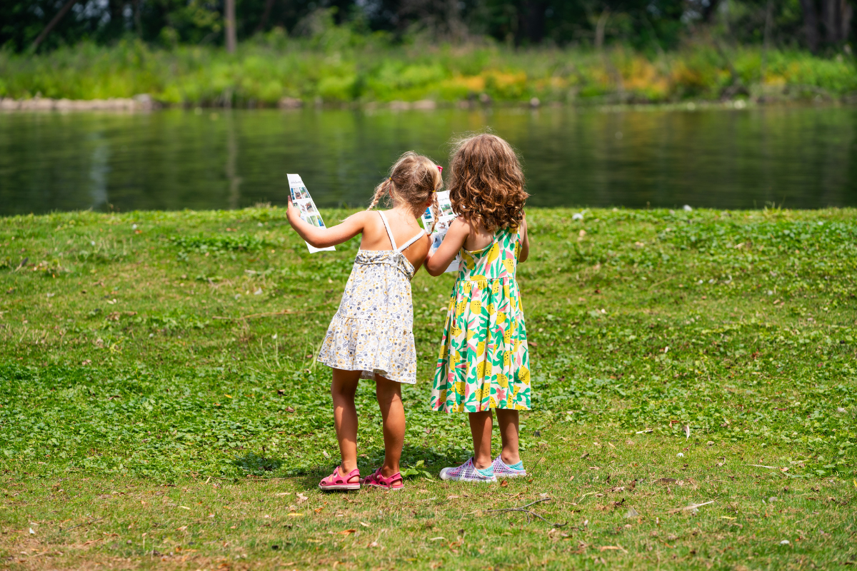 Two young girls stand on a grassy field near a calm pond, each holding an illustrated activity sheet, appearing to explore or participate in an outdoor scavenger hunt on a sunny day.