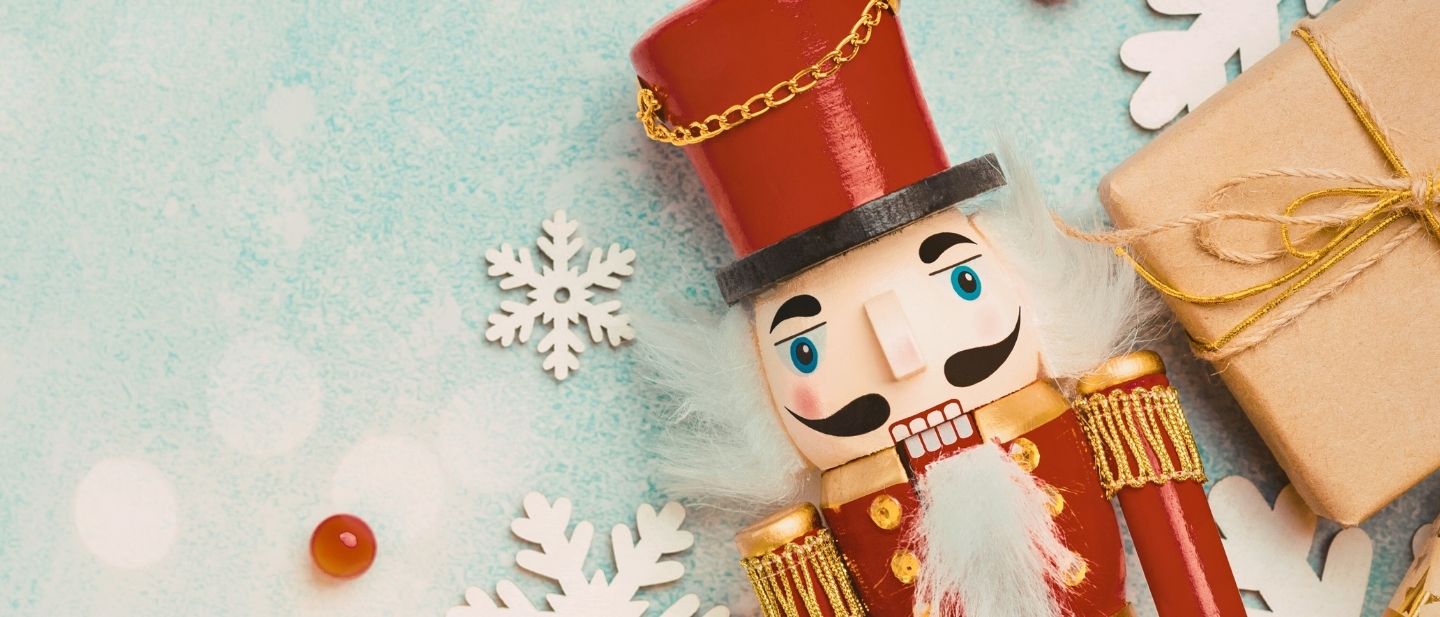 A classic red Nutcracker figurine rests on a light blue winter background surrounded by white snowflake cutouts and a wrapped gift, evoking the Nutcracker Tea holiday theme.