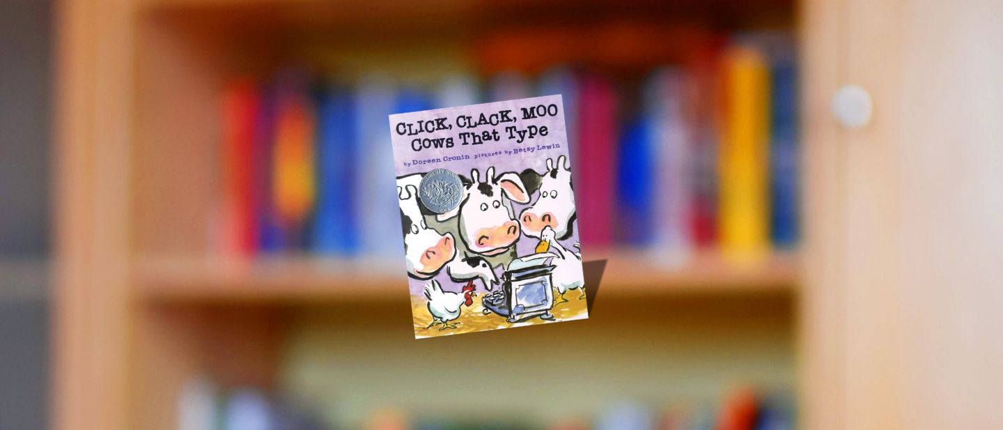 The cover of Click, Clack, Moo: Cows That Type by Doreen Cronin displayed against a blurred colorful bookshelf in the Jensen/Kahn Classrooms at Ford House.