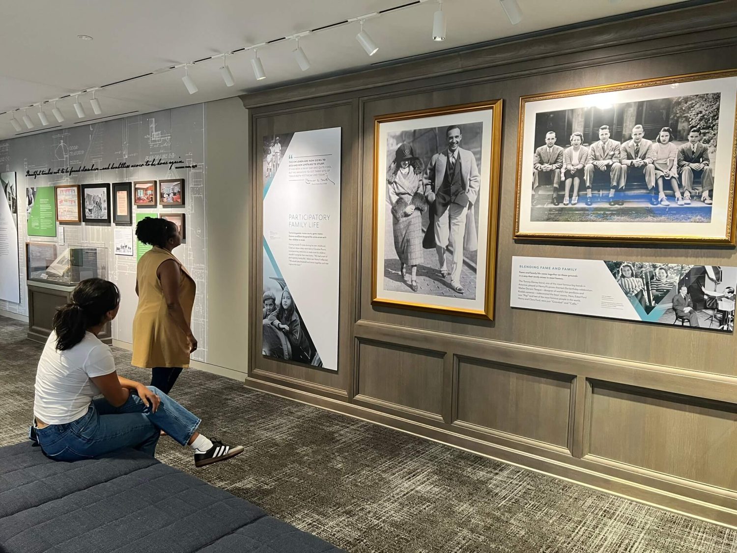 Two visitors examine an exhibition display about Ford family life. The wall features multiple framed black and white photographs of family members spanning different eras, alongside interpretive text panels about participatory family life. The gallery space has professional museum lighting and wood-paneled walls.