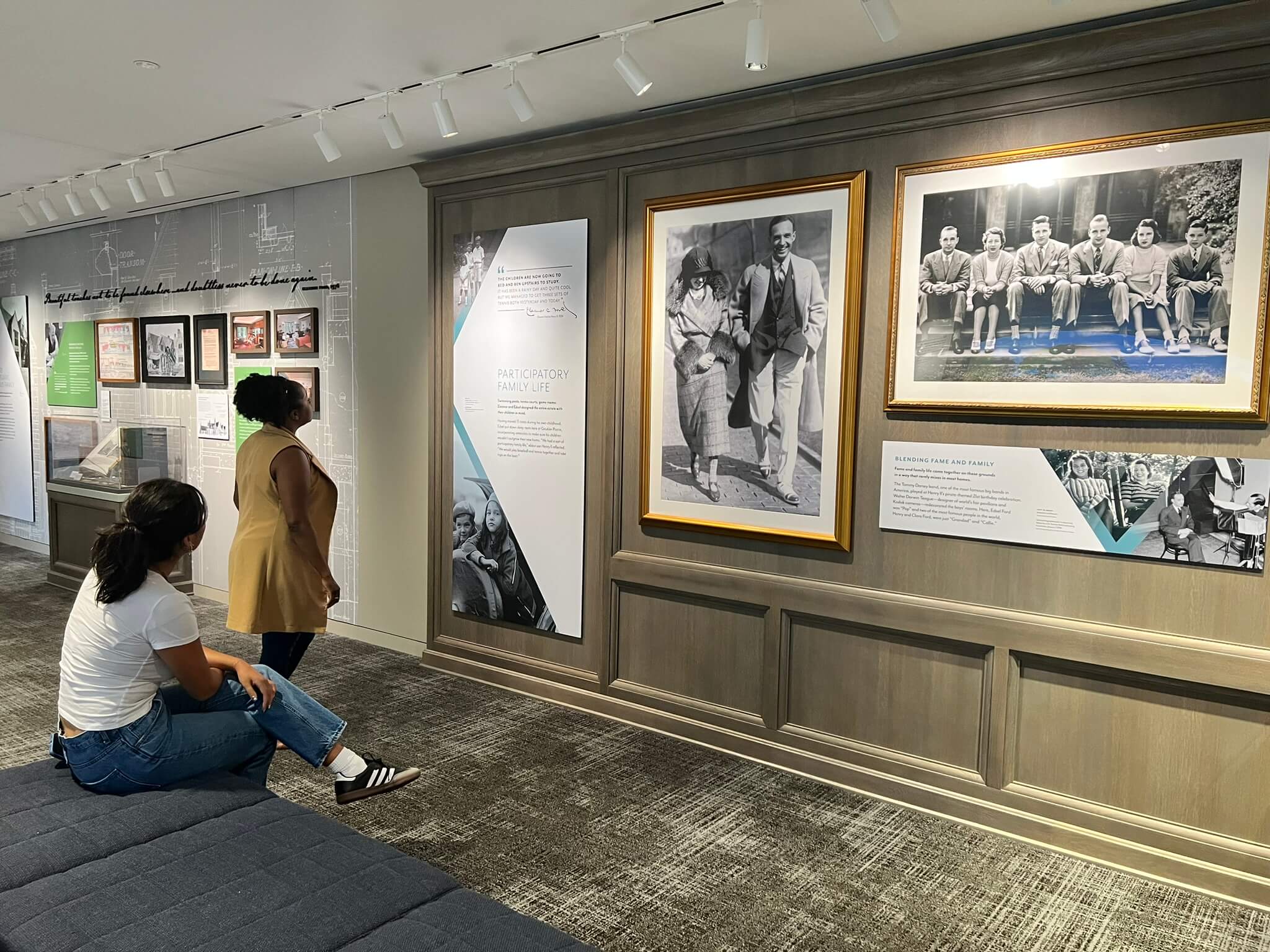 Two visitors examine an exhibition display about Ford family life. The wall features multiple framed black and white photographs of family members spanning different eras, alongside interpretive text panels about participatory family life. The gallery space has professional museum lighting and wood-paneled walls.