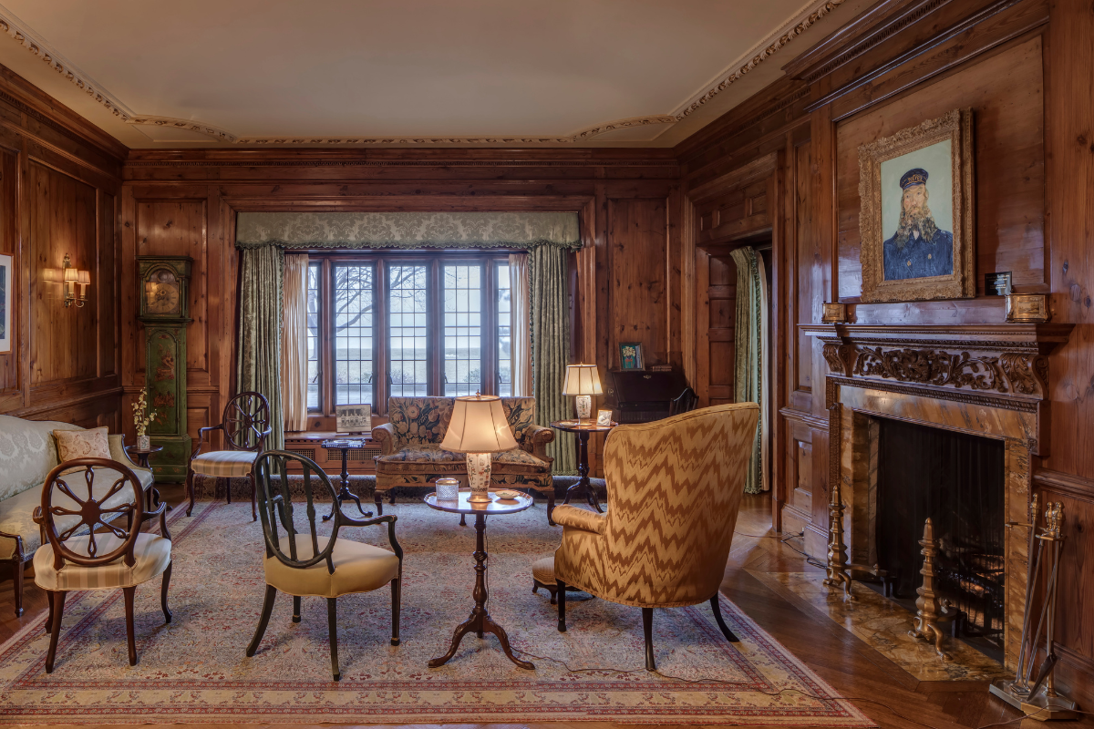 An elegantly appointed historic morning room features rich wood paneling, an ornate fireplace with carved mantelpiece, and period furnishings including upholstered chairs and side tables. Large multi-paned windows with silk drapery overlook the grounds. The room's coffered ceiling, original artwork, and warm lighting preserve the refined aesthetic of early twentieth-century domestic luxury.