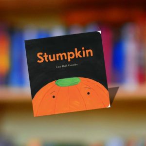The cover of Stumpkin by Lucy Ruth Cummins displayed against a blurred colorful bookshelf in the Jensen/Kahn Classrooms at Ford House.