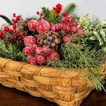 Woven splint basket of evergreen sprigs, frosted red berries and pinecones on a dark wood table.