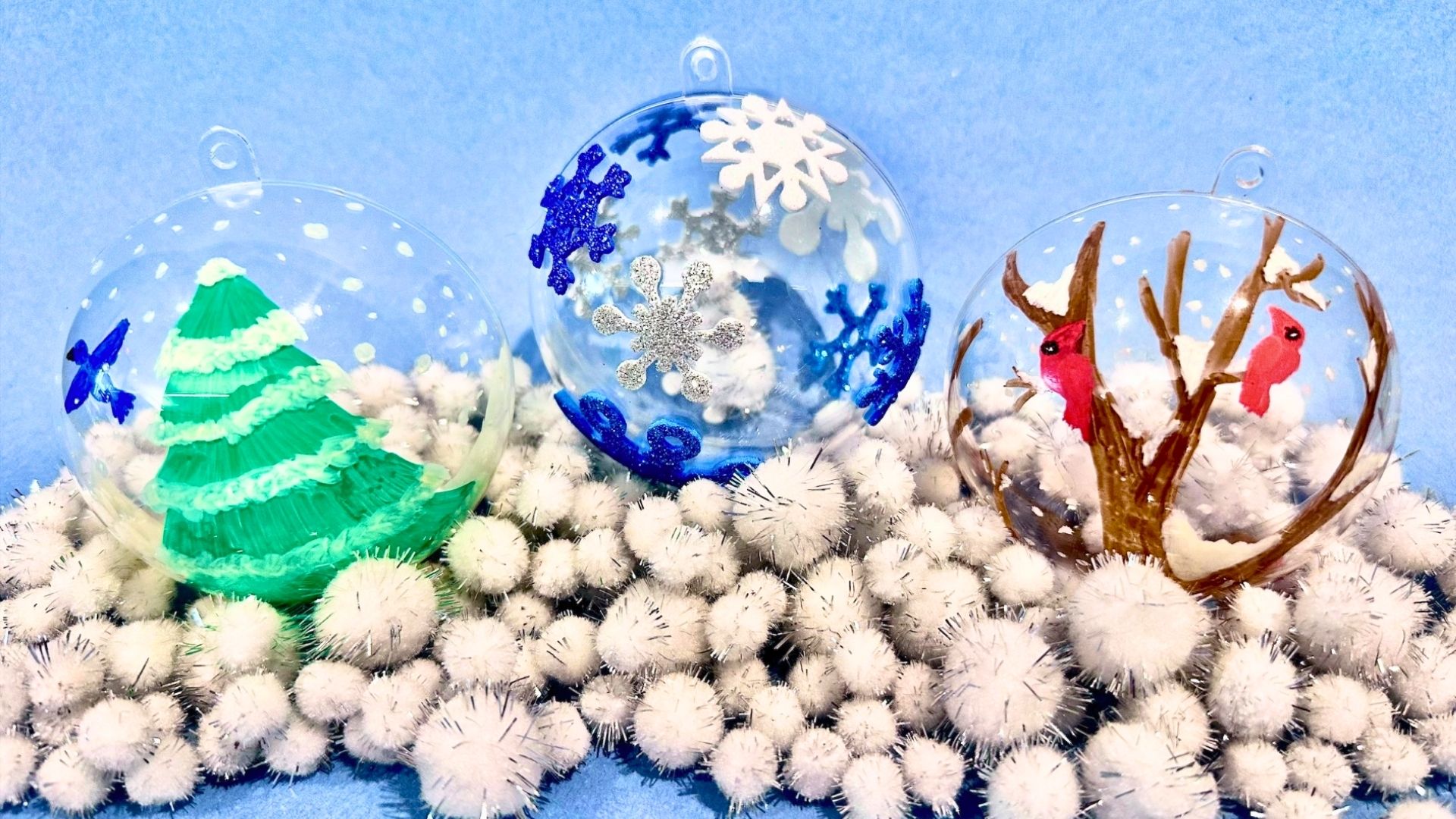 Three hand-painted clear glass ball ornaments, a tree, snowflakes, and cardinals, resting on white pom-pom snow.