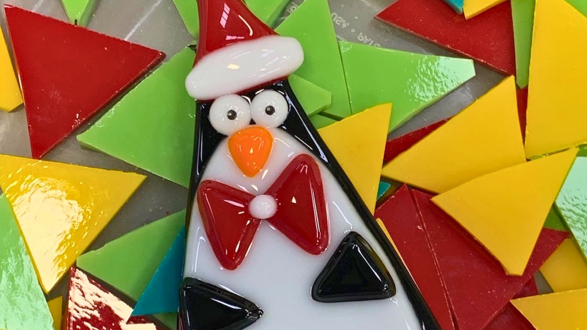 Fused glass penguin ornament with a red Santa hat and red bow tie, set on colorful glass triangle pieces.