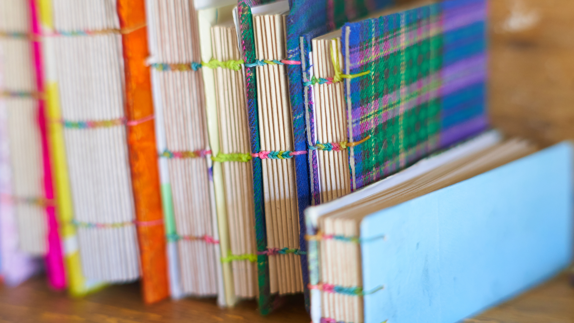 Row of handmade hardcover books with colorful covers, spines bound with visible cross-stitch thread, on a wood shelf.